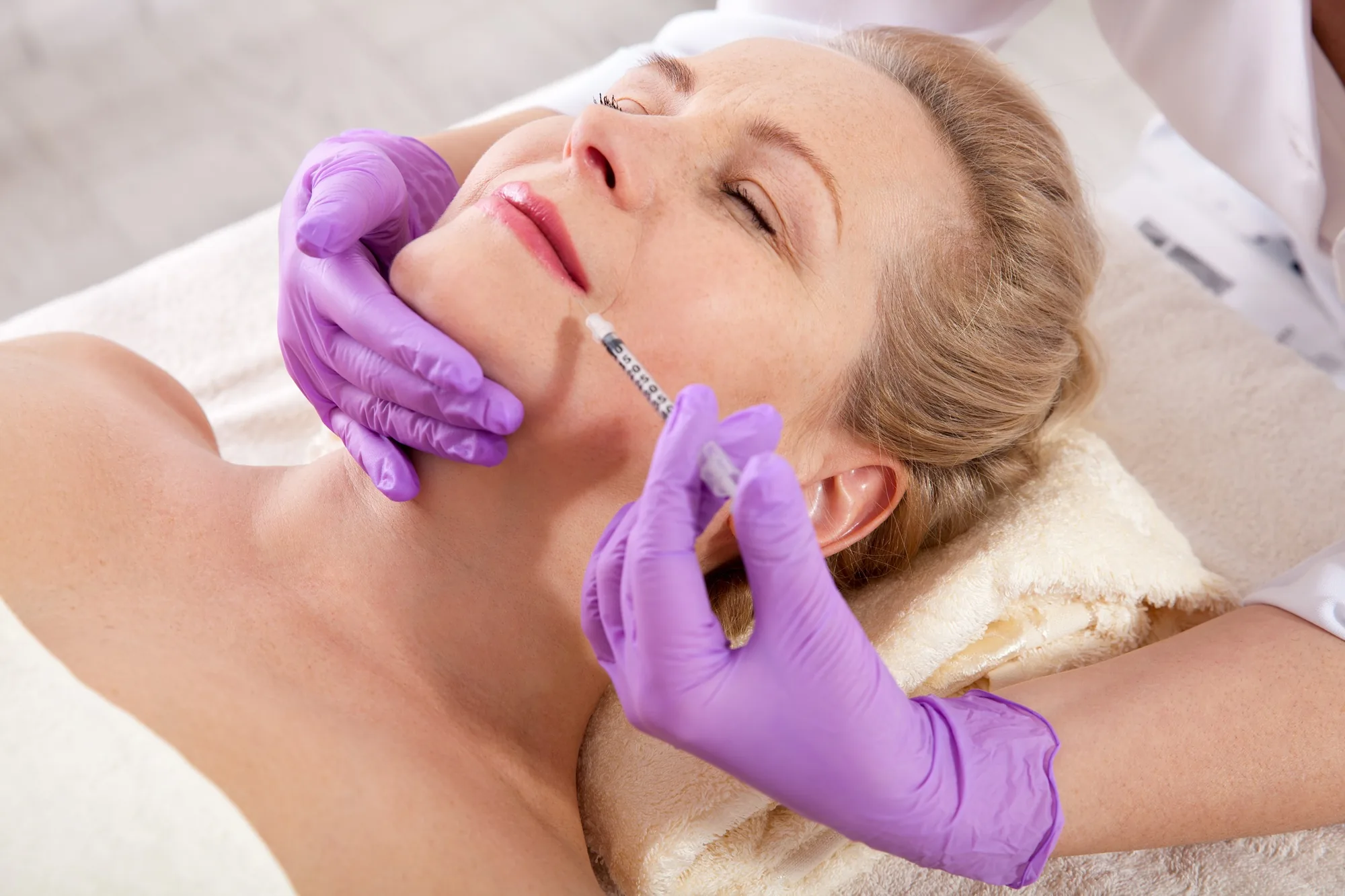 Doctor wearing purple gloves injecting a middle-aged woman's chin area